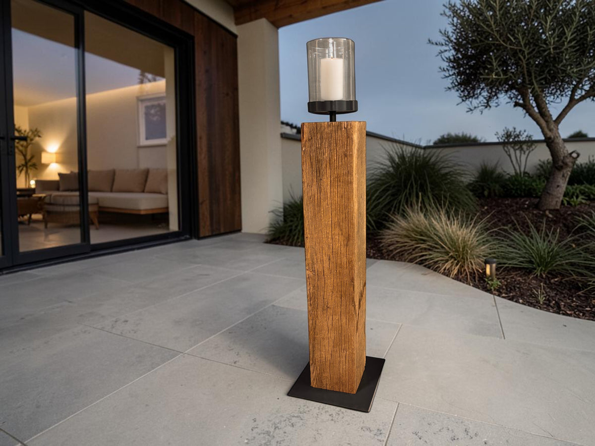 Windlichtsäule aus antiker Eiche auf einer geschützten Terrasse in früher Dämmerung; inszenierter Hintergrund und ergänzte Kerze mit Flamme.